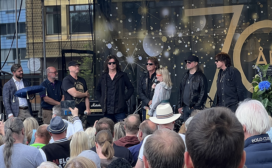 På en scen står alla fem medlemmar från bandet Europe, tillsammans med styrelseordförande Oskar Weinmar, konferencier Josefin Sundström och Roland Storm. Nedanför scenen syns en publik. 